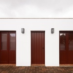 Červené vstupní dveře, které kontrastují s neutrální fasádou a dávají každému domu jasně čitelný bod vstupu. Barevný akcent dveří není pouze estetický – odkazuje na místní tradici používání výrazných tónů pro oživení jednoduchých objemů. Foto: Carmo Oliveira