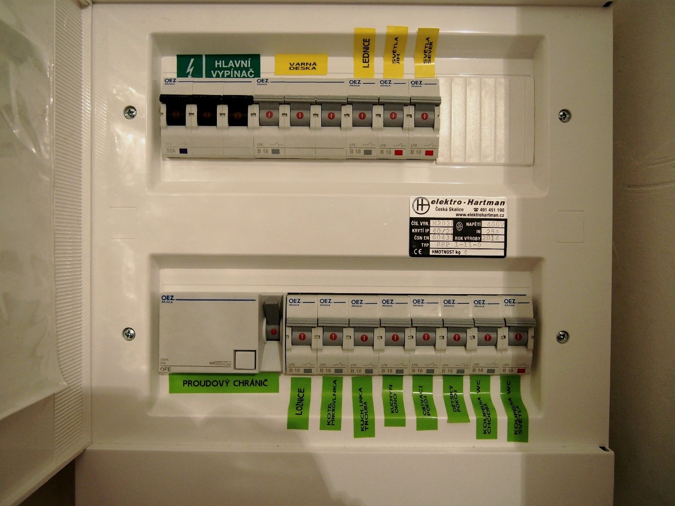 Pohled do bytové rozvodné skříně. 1 řádek zleva: Hlavní vypínač bytový třífázový (3x 32A – musí odpovídat hlavnímu jističi na patře), třífázový jistič pro varnou desku, jistič na zásuvku lednice a dva světelné okruhy. 2. řádek zleva: Proudový chránič, 8 zásuvkových okruhů (pro každou místnost zvlášť – obývací pokoj, 2x ložnice, koupelna zásuvky, koupelna světla, tři okruhy pro kuchyň). Skříň má revizní štítek o tom, že byla odborně sestavena.