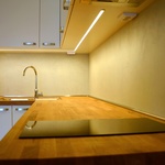 Detail kuchyňské desky z masivu, na kterém je patrná jak indukční dvouplotýnka, tak i dřez a LED podsvícení. Zásuvky a vypínač jsou umístěny do zdvojeného dna vrchních skříněk kuchyňské linky
